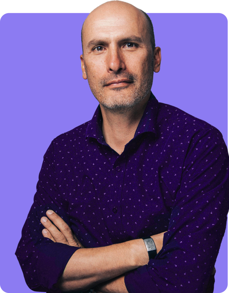 Man with a shaved head wearing a purple shirt with a pattern, arms crossed in front of a solid background.