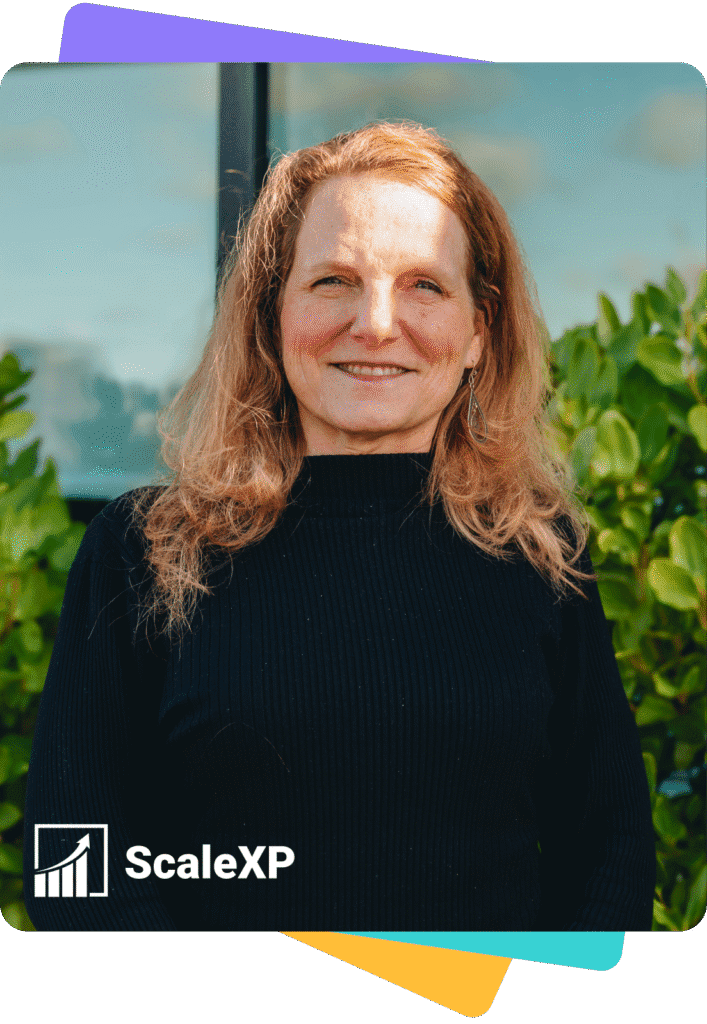 Woman with long hair wearing a black sweater, standing in front of greenery with a ScaleXP logo in the corner.